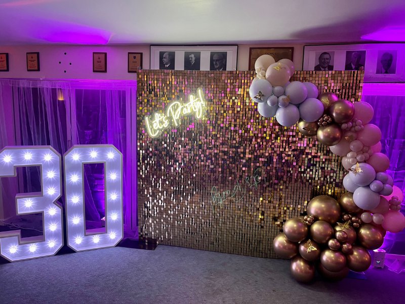 Gold sequin shimmer wall backdrop with Let's Party neon sign and balloon garland event decor