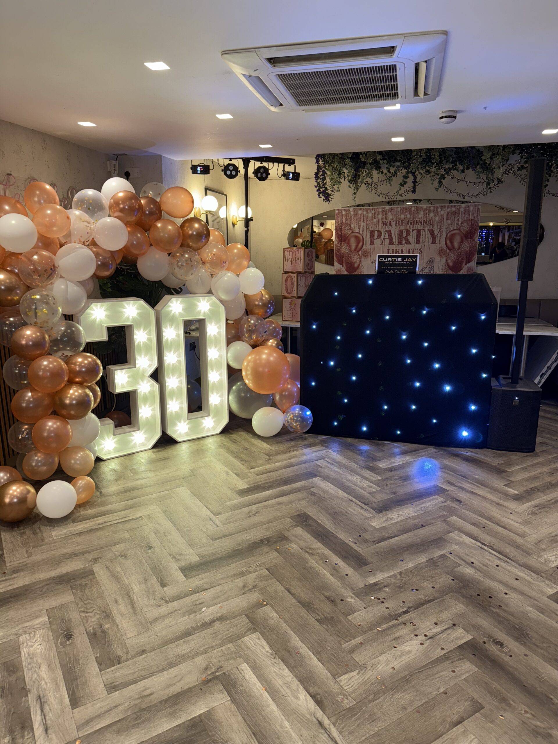 Light-up 30 numbers with rose gold and white balloon garlands beside a starlit DJ booth