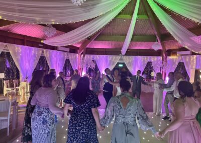 Wedding guests dancing in a circle under fairy-light drapes and pink venue lighting at The Ravenswood