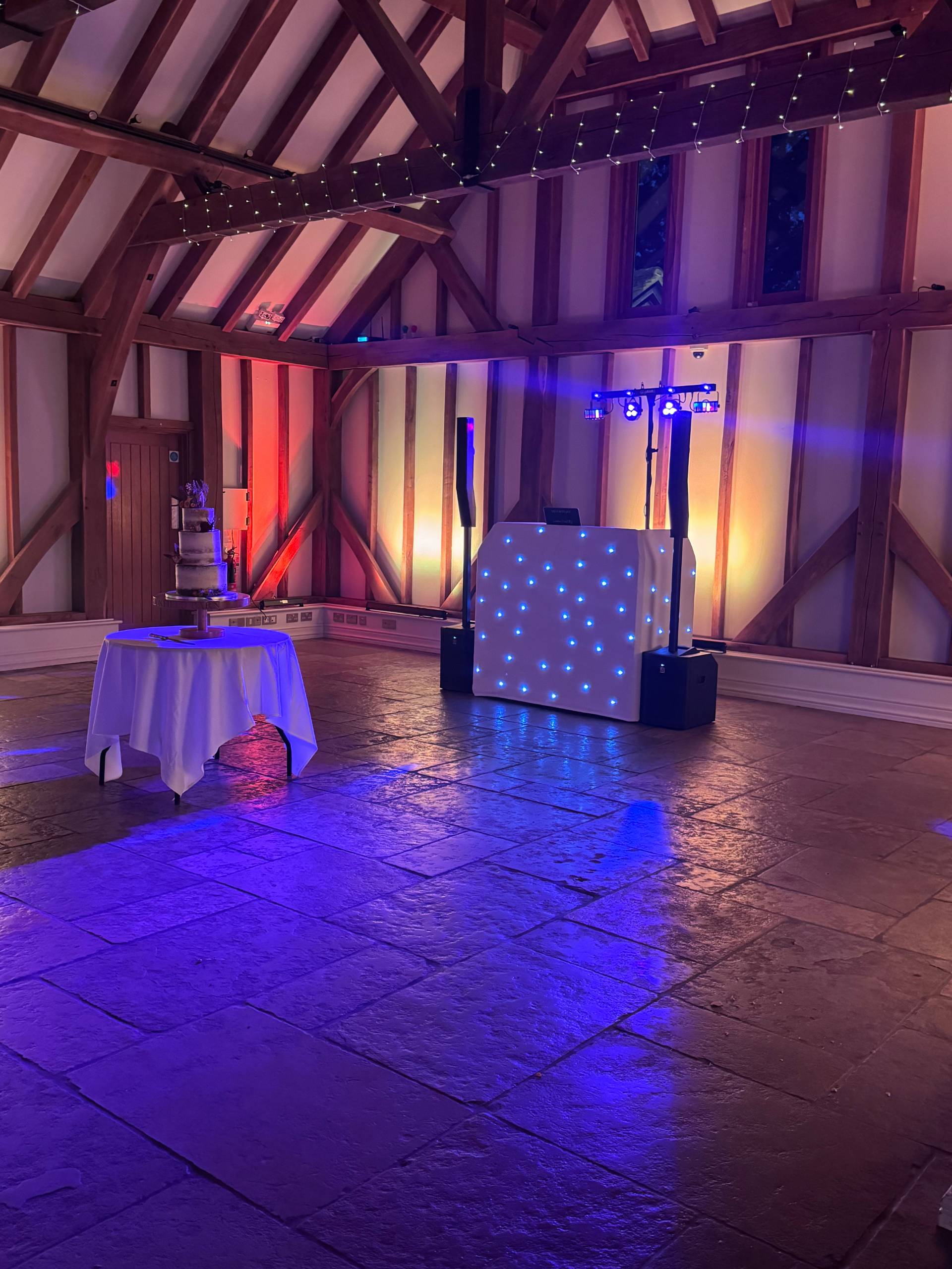 Wedding breakfast set-up with three-tier cake and starlit DJ booth in a beamed barn venue
